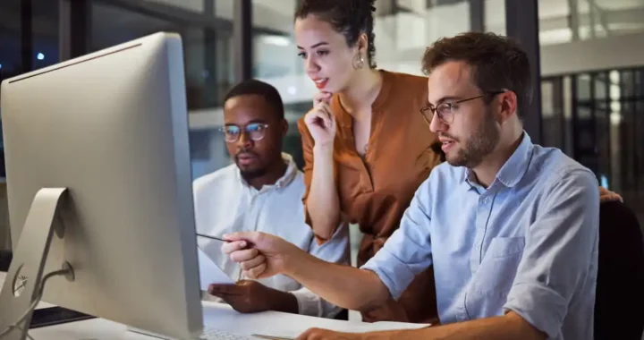 Equipe de três pessoas colaborando em frente a um computador, discutindo um fluxo de trabalho compartilhado