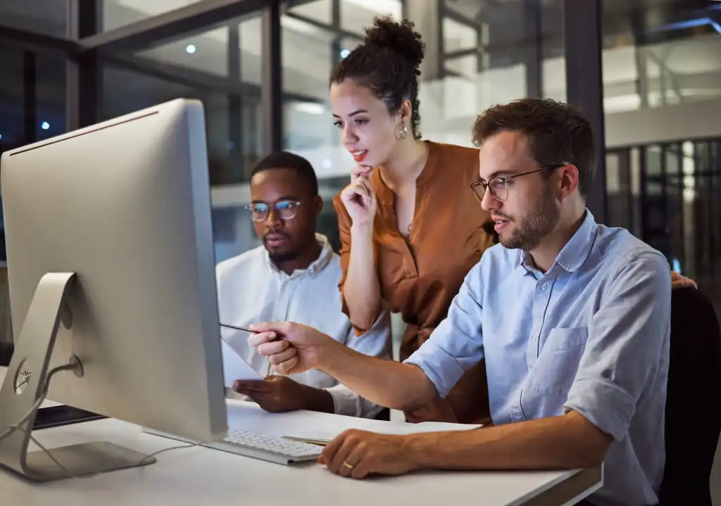 Equipe de três pessoas colaborando em frente a um computador, discutindo um fluxo de trabalho compartilhado